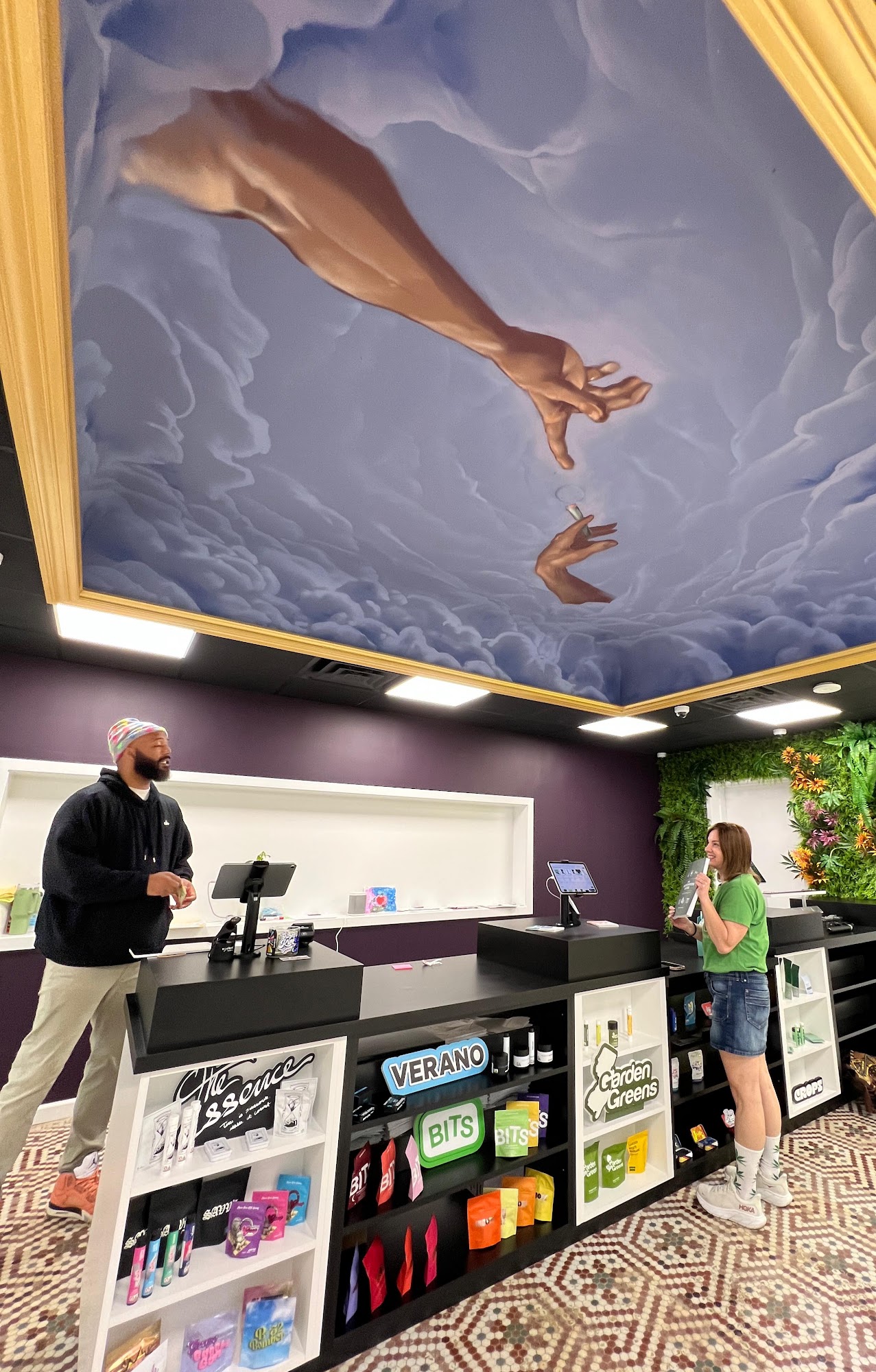 A budtender helping a smiling customer beneath the ceiling mural at Hamm & Chaz, Jersey City